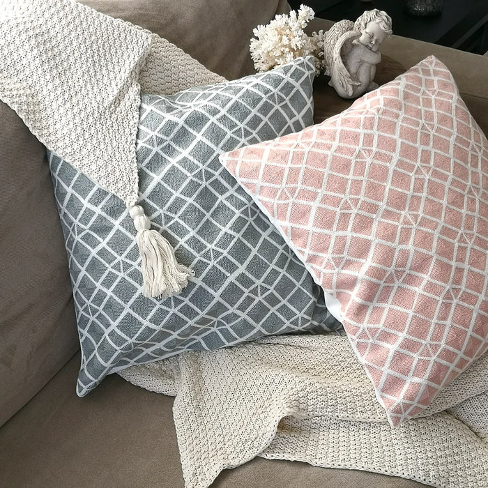Two square throw pillows, one grey and one pink, both with a woven white diamond pattern, resting on a beige textured throw blanket on a couch. A cherub statue and white decorative object are behind the pillows.