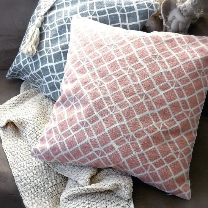 Two square throw pillows, one grey and one pink, both with a woven white diamond pattern, resting on a beige textured throw blanket on a couch. A cherub statue and white decorative object are behind the pillows.