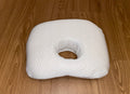 A white baby comfort pillow for head support, featuring a soft, textured cover and a central hole to help prevent flat head, shown on a light wood floor.