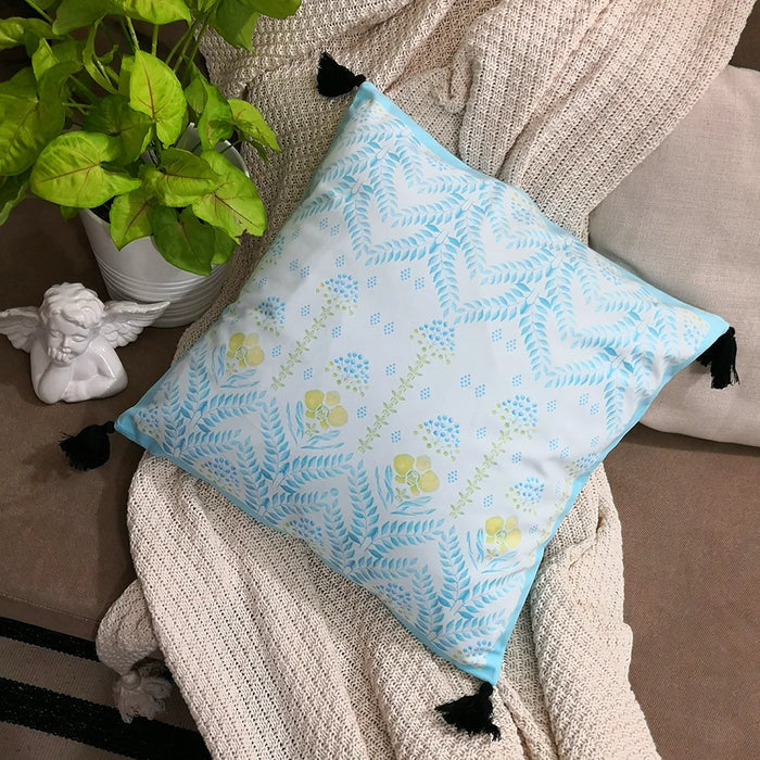 A square white pillow with light blue, yellow, and green floral and leaf patterns, featuring black tassels, rests on a light beige textured blanket next to a green plant and white cherub statue