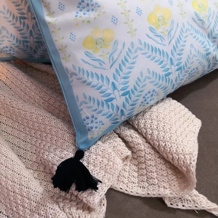 Close-up of a white pillow with light blue, yellow, and green patterns, showing a black tassel and the texture of a beige knitted blanket