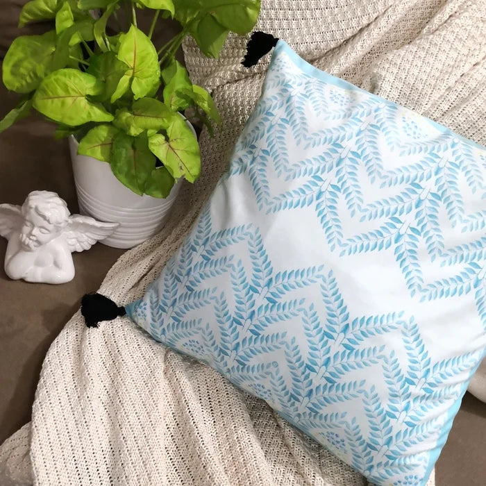 A square white pillow with a light blue and green chevron leaf pattern, featuring black tassels, rests on a light beige textured blanket next to a green plant and white cherub statue