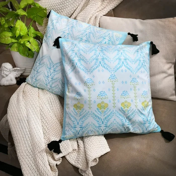 Two square white pillows with light blue and green patterns, one floral and one chevron, featuring black tassels, rest on a light beige textured blanket next to a green plant and white cherub statue.