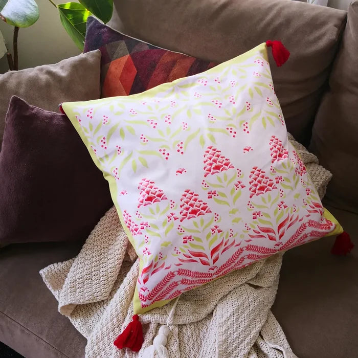 Light colored pillow with red floral and berry print and red tassels on a couch with other pillows and a textured blanket.