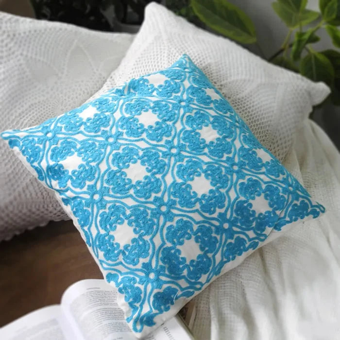 White square pillow with blue embroidered floral pattern on a couch with open book, white textured blanket, and plant in the background