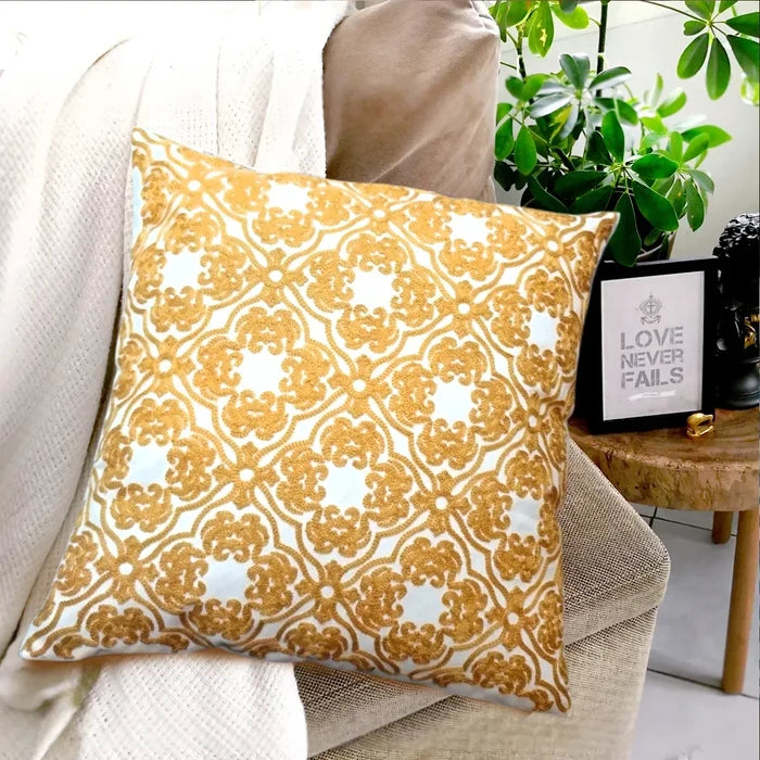 A white square pillow with golden embroidered pattern on a couch, with blanket, plants, and framed art in background.