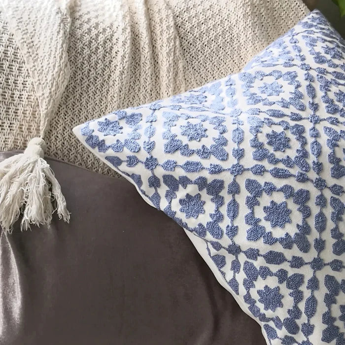 A white throw pillow with intricate blue embroidered floral patterns rests on a gray velvet pillow and a light beige knitted blanket. A white tassel is visible